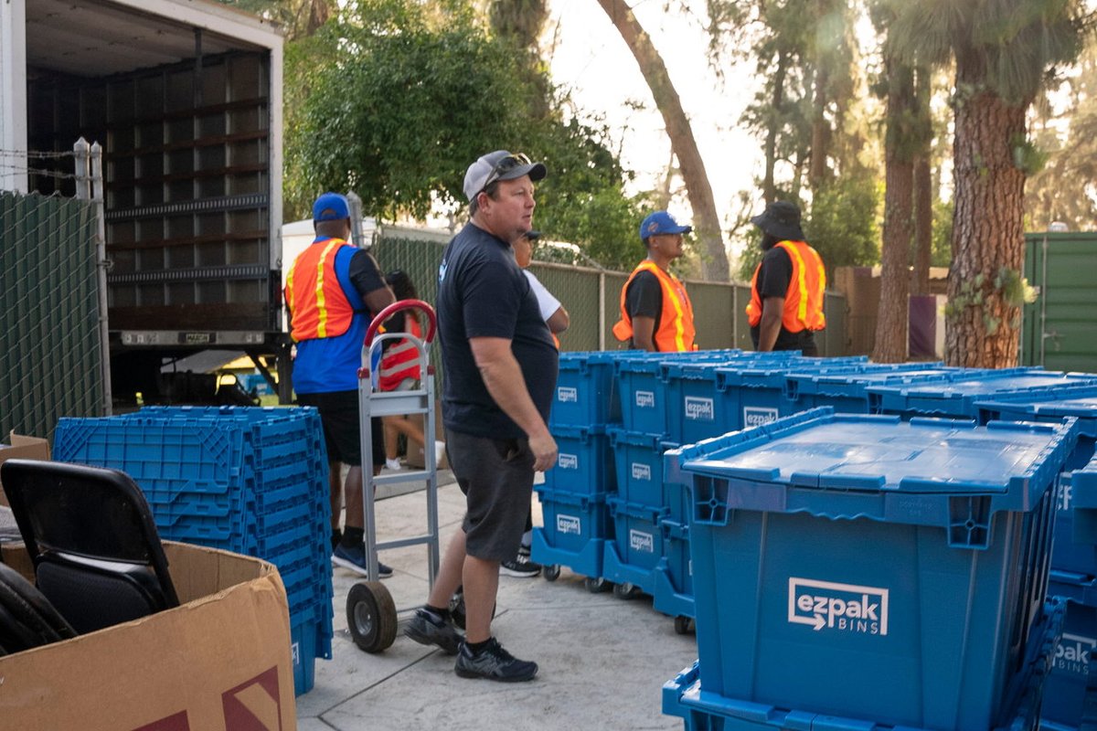 A crew loads stacked blue EZPak Bins moving crates near an open box truck in a sunlit outdoor lot, with a hand truck and boxes nearby