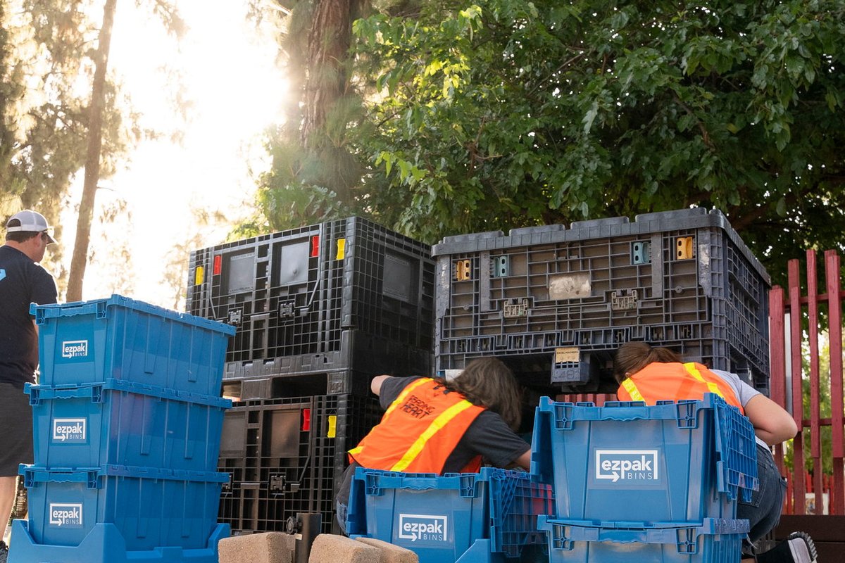 A crew loads blue EZPak Bins moving crates near large storage containers outdoors