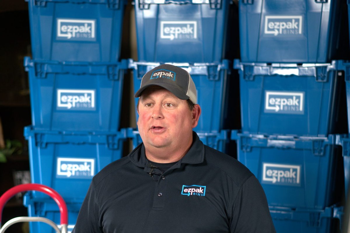 A man in an EZPak Bins branded polo and cap stands with a hand truck in front of stacked blue moving bins
