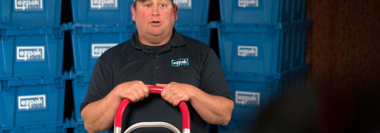 A man in an EZPak Bins ballcap and polo grips a red hand truck in a warehouse with shelves of stacked blue moving bins behind him