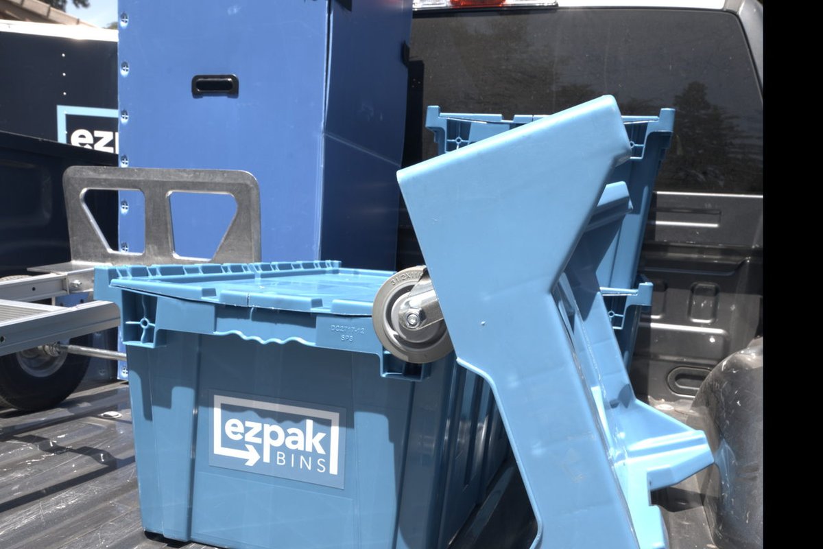 Close-up of a blue EZPak Bins moving bin with its lid propped open next to a folded hand dolly in a pickup truck bed