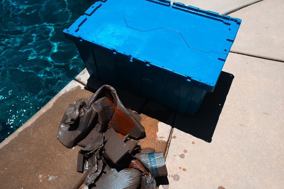 A dry blue EZPak Bins moving bin sits beside a pool next to soggy, falling-apart wet cardboard boxes, showing durability versus cardboard