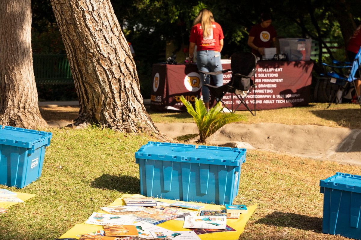 Blue ezpak bins sit on a grassy area lining a yellow tarp covered with books at an outdoor community event, with people at an information booth in the background under trees