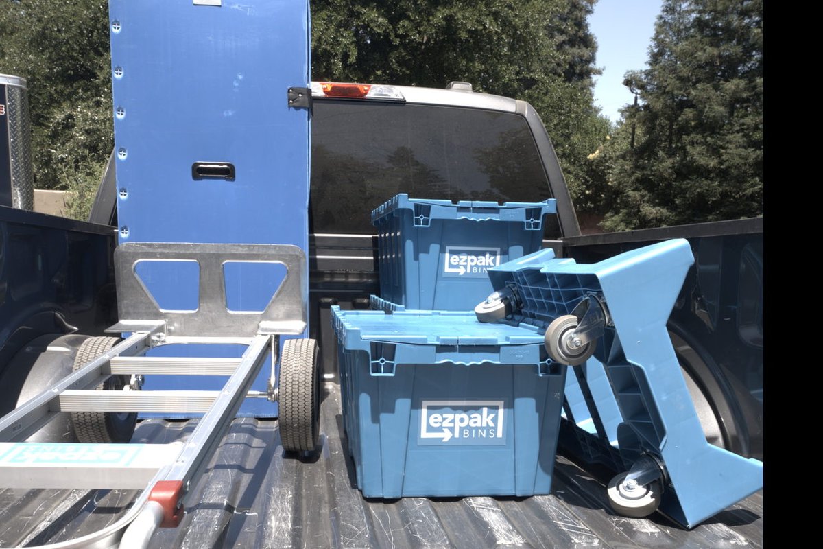 Two stacked blue EZPak Bins moving crates and a red-handled hand truck loaded in the bed of a pickup truck parked under trees