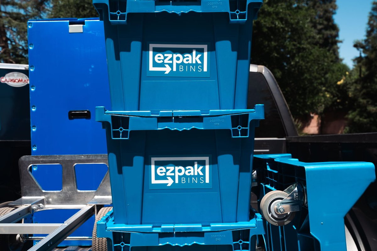 Three blue stackable EZPak Bins moving crates and a hand dolly stacked in the bed of a pickup truck, parked outdoors under trees
