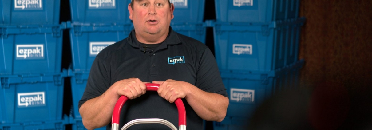 A man in an EZPak Bins branded cap and polo grips a red-handled hand truck in front of stacked blue moving bins