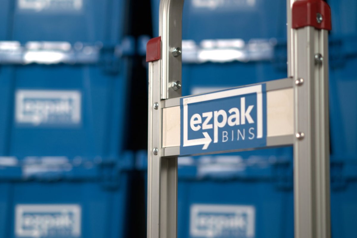 A close-up of an EZPak Bins branded hand truck with a red handle in front of rows of stacked blue moving bins in a warehouse