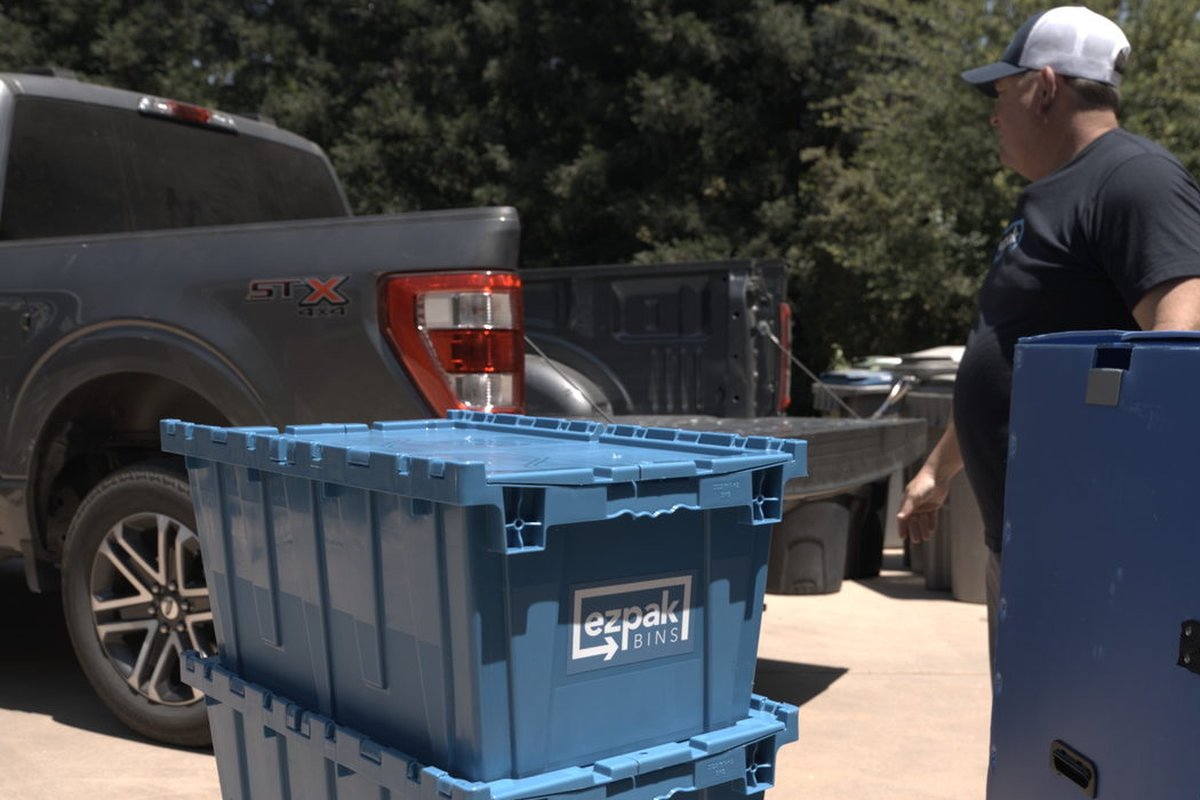 Stacked blue EZPak Bins moving crates beside a pickup truck outdoors, with a man in a cap nearby