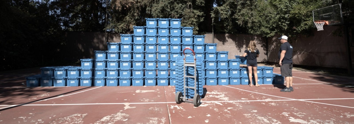 A hand truck stands ready on an outdoor court in front of a large pyramid-shaped stack of blue EZPak Bins moving crates, with two people beside it