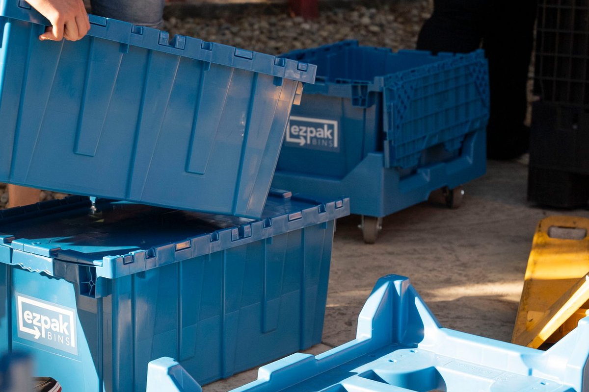 A person lifts the lid of a stacked blue EZPak Bins moving bin next to an empty rolling dolly cart outdoors