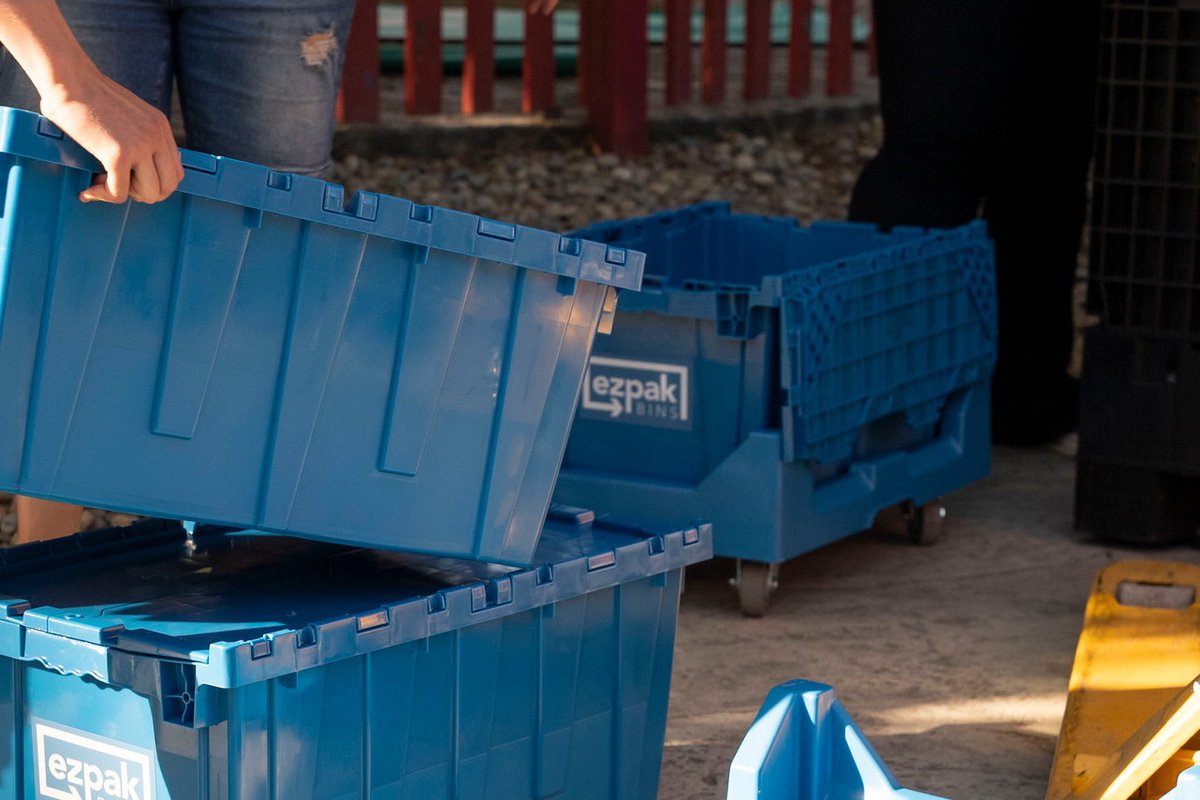 A person lifts the lid off a stacked blue EZPak Bins moving crate next to an empty rolling dolly, outdoors by a red fence