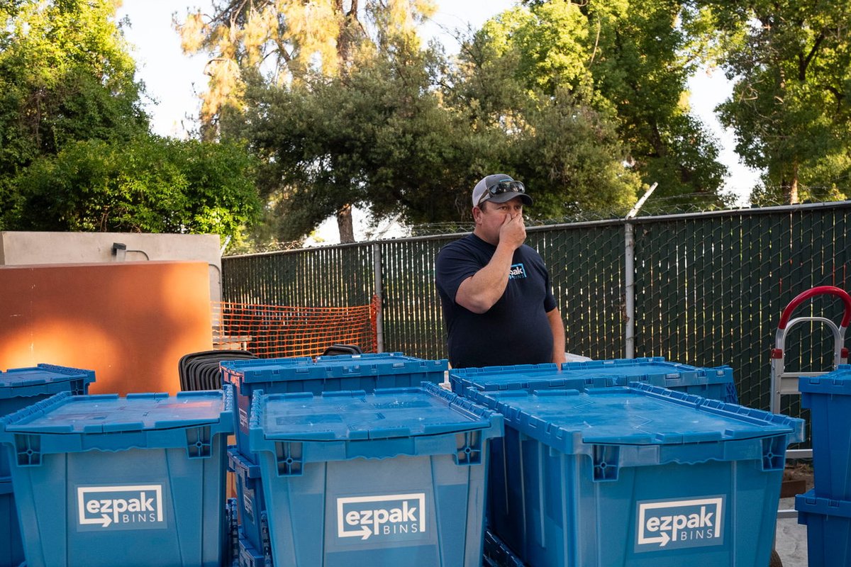 A man in a cap and dark shirt stands behind rows of stacked blue EZPak Bins moving crates on dollies in an outdoor lot