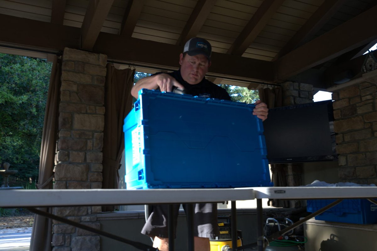 A man in a cap carries a large blue plastic moving bin through a covered patio area next to a stone wall