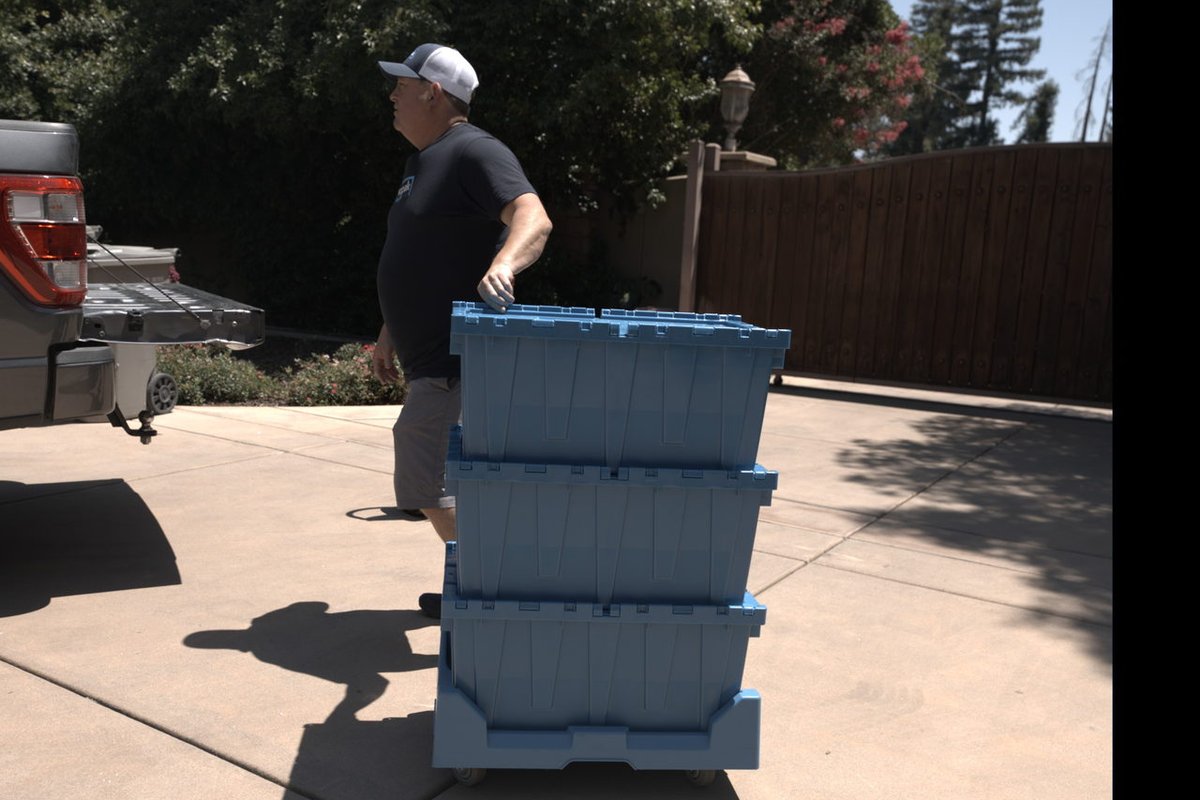 A man in a cap and dark shirt carrying a stack of blue moving bins on a residential driveway, with his shadow cast on the pavement and a tree and house in the background