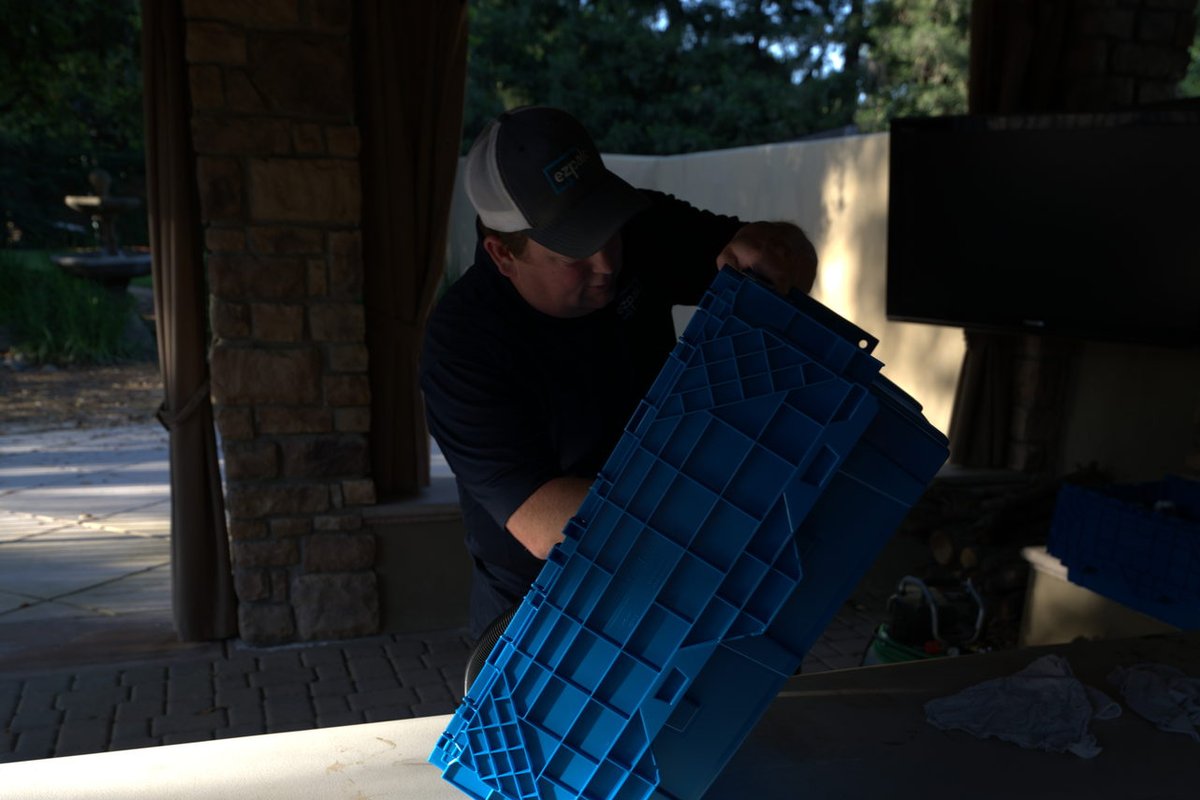 A man in a cap carries a stack of blue EZPak Bins moving bins past a stone pillar near a house entryway