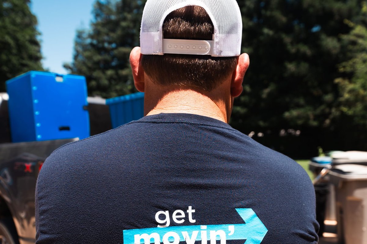 A man wearing an EZPak Bins Get Movin branded shirt stands near blue moving bins and a truck