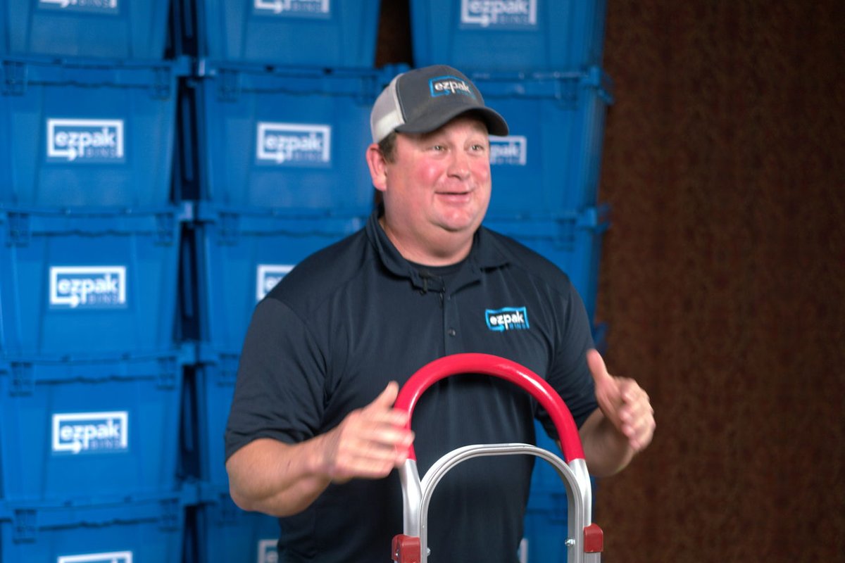 A man in a cap and polo holds a red and silver EZPak Bins branded hand truck in front of stacks of blue moving bins