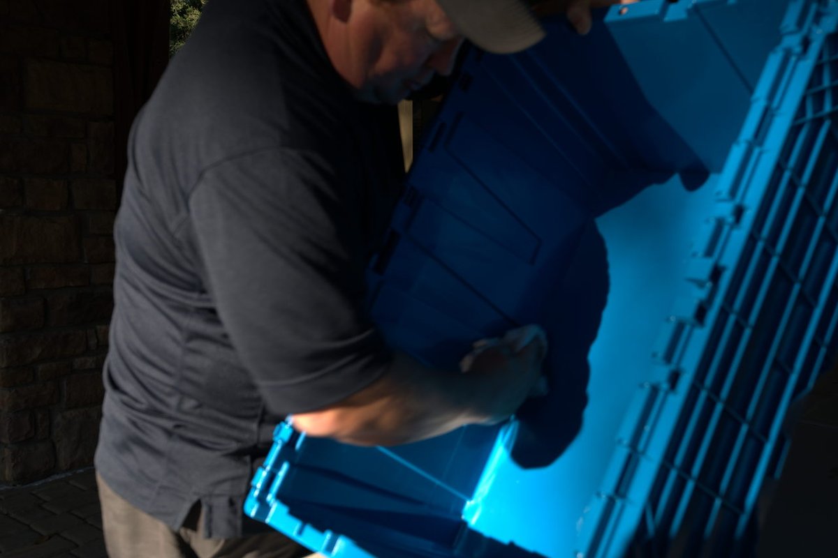 Close-up of a man in a cap leaning over and reaching into an empty blue plastic moving bin outdoors