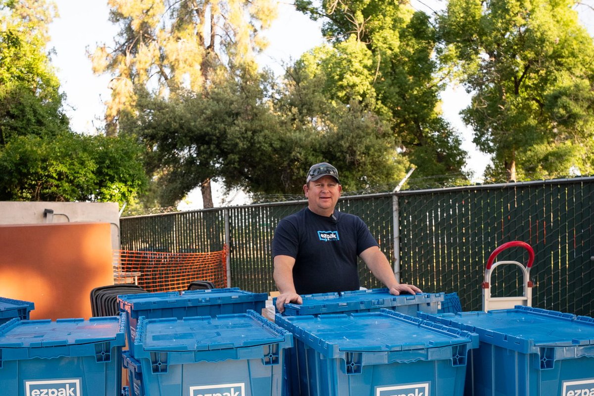 A smiling man in a cap and EZPak Bins polo stands with his hands on stacks of blue EZPak Bins moving crates in an outdoor yard