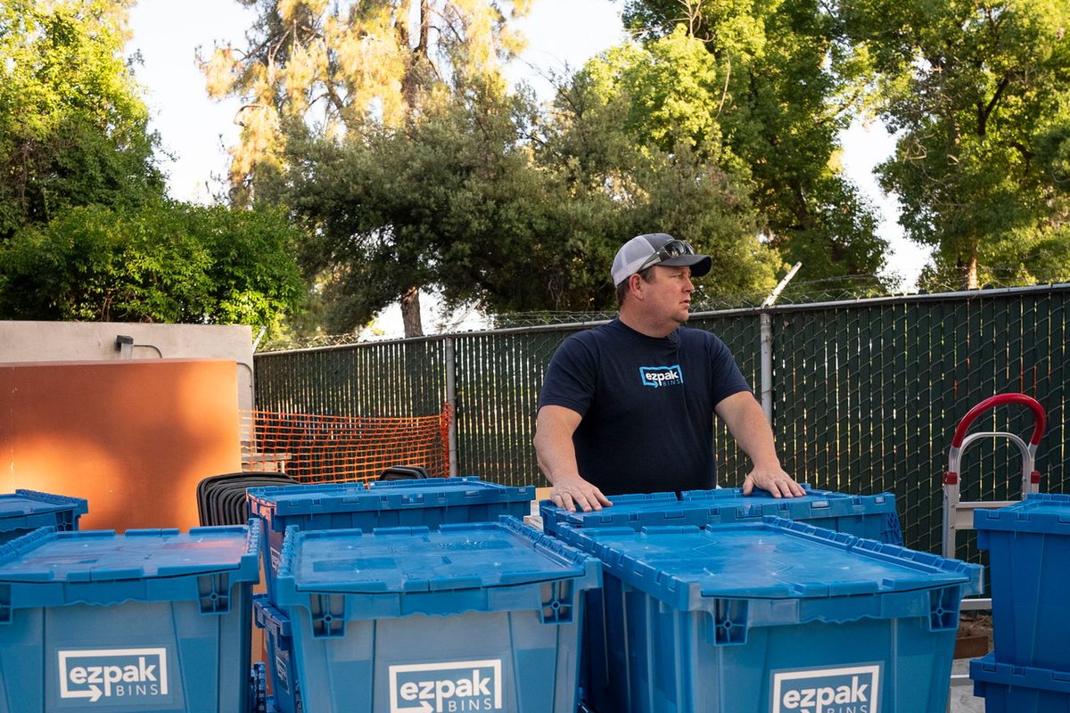 A man in an EZPak shirt and cap stands behind stacked blue EZPak Bins moving crates in an outdoor yard