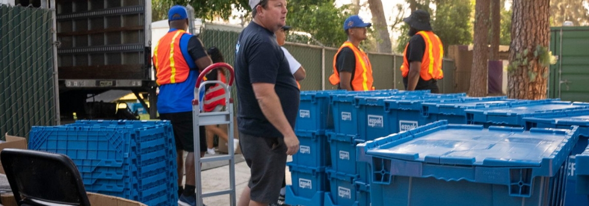 A crew unloads stacked blue EZPak Bins moving crates from an open box truck in an outdoor lot, with a hand truck nearby