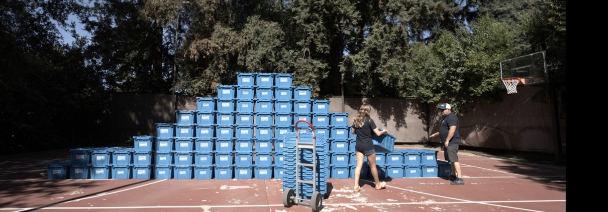A man and a woman carry a blue moving bin toward a hand truck on a backyard basketball court, with a large pyramid-shaped stack of bins behind them