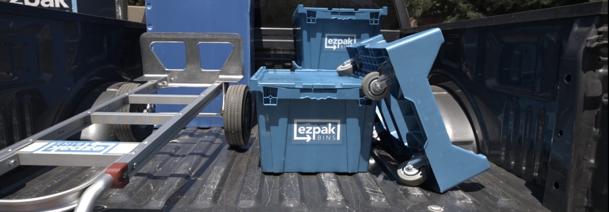 Two blue EZPak Bins moving crates and a folded hand truck in the bed of a pickup truck, with an EZPak-branded box truck parked behind