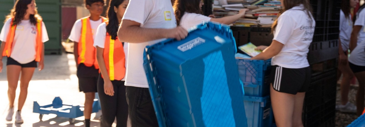 A man carries an empty blue EZPak Bins moving crate outdoors as a line of helpers sorts books nearby