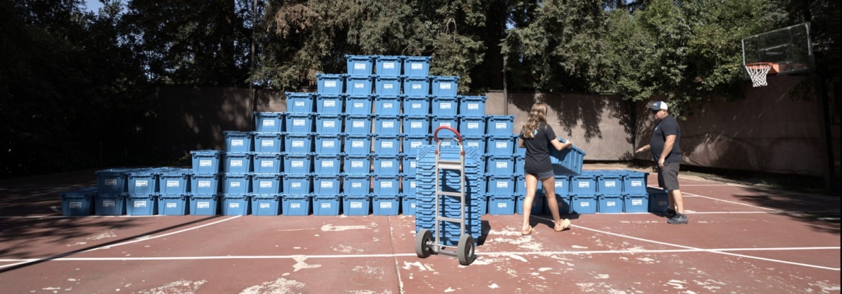 A man and a woman carry a blue EZPak Bins moving crate toward a hand truck on an outdoor court stacked with more bins