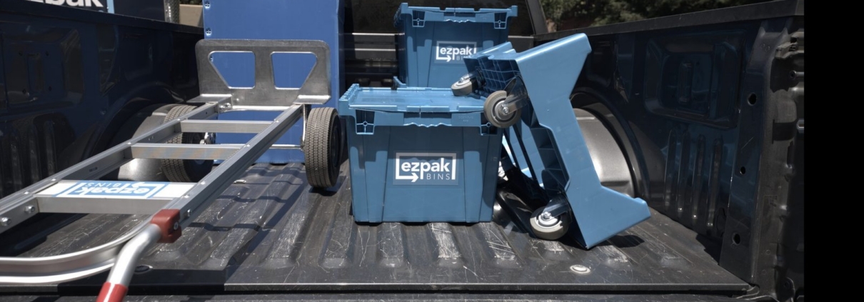 Two blue EZPak Bins moving crates and a folded blue hand truck sit in the bed of a pickup truck, with a large ezpak-branded box truck parked behind it and trees in the background
