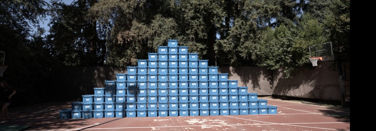 A tall pyramid-shaped stack of blue EZPak Bins moving bins fills a backyard basketball court surrounded by trees