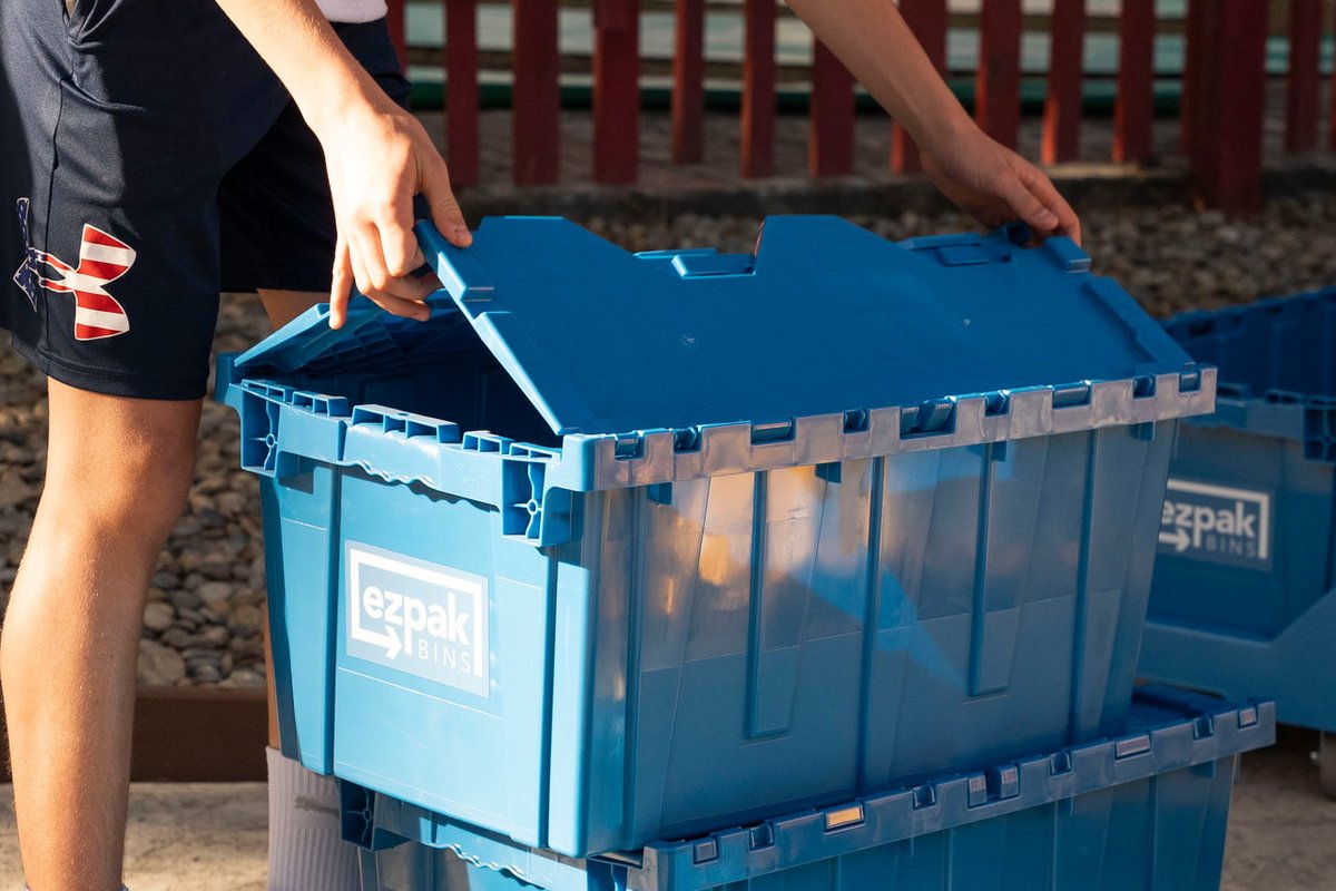 A person opens the lid of a stacked pair of blue EZPak Bins moving crates outdoors by a red fence