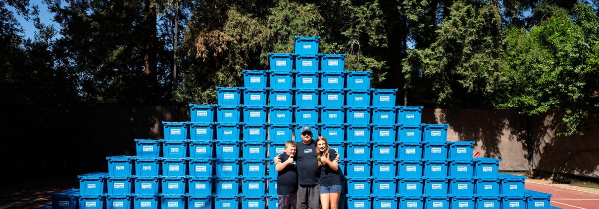 A family poses in front of a massive full-width pyramid of blue EZPak Bins moving bins on an outdoor court, showing the large supply of packing bins on hand
