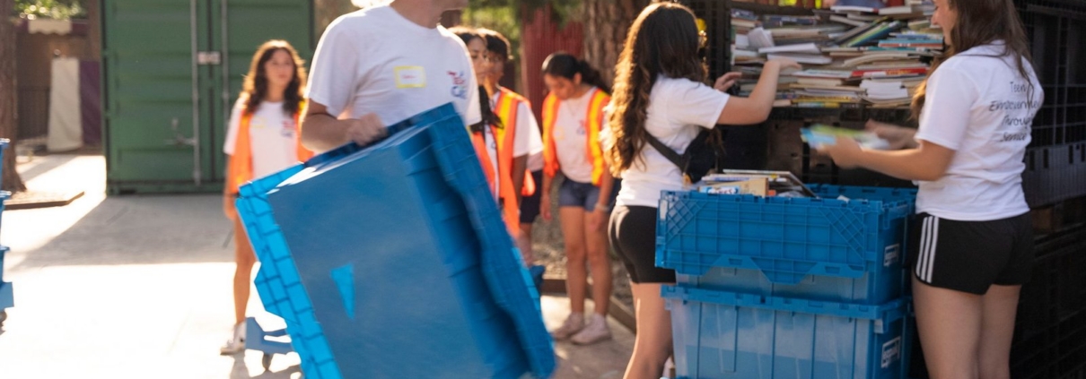 A man carries an empty blue EZPak moving bin outdoors while people in the background sort items into stacked blue bins