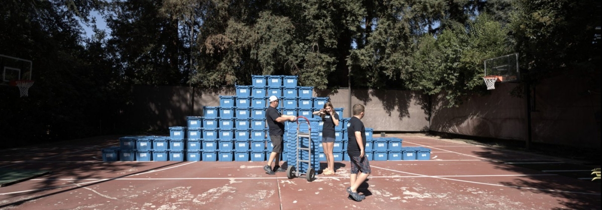 Workers gather around a hand truck loaded with blue EZPak Bins moving crates on an outdoor court stacked with more bins