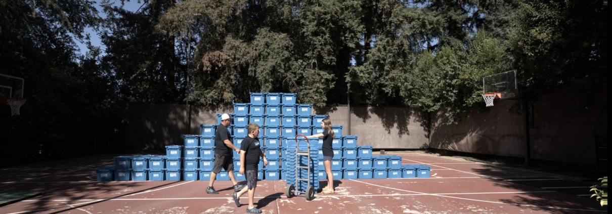Workers on an outdoor court beside a large pyramid-shaped stack of blue EZPak Bins moving crates, with one pushing a hand truck loaded with more bins