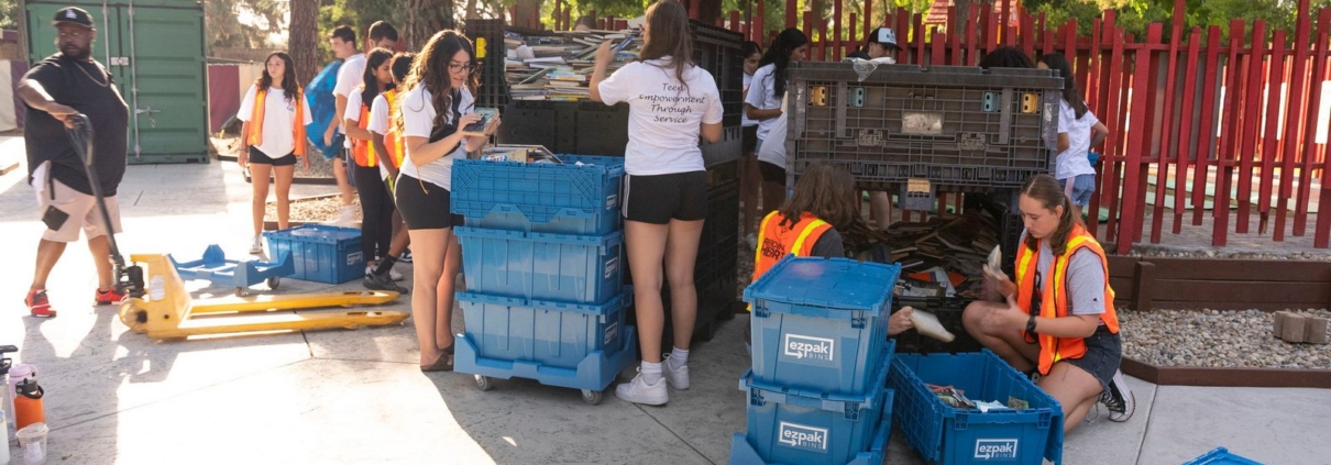 A group sorts books and supplies into blue EZPak Bins moving crates at an outdoor community event near Woodward Park