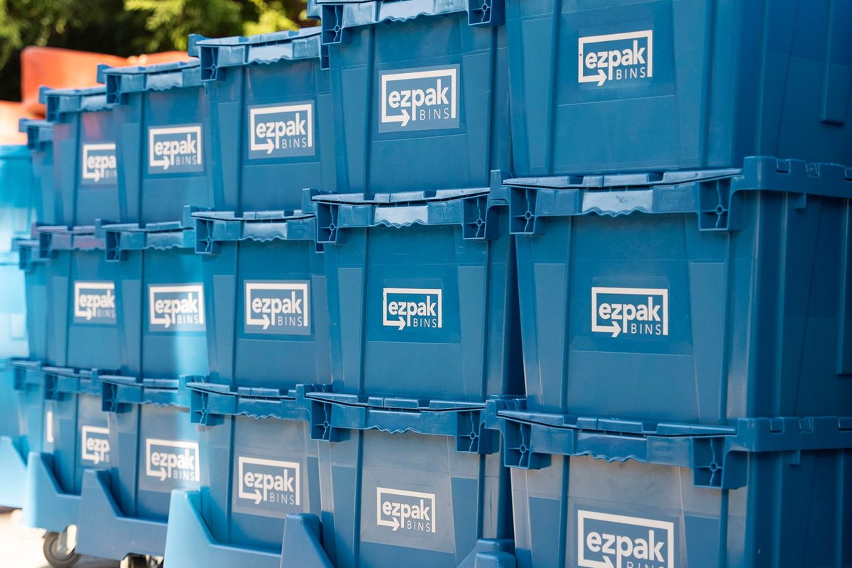 Rows of stacked blue EZPak Bins moving crates on wheeled dollies lined up on a concrete driveway with trees behind