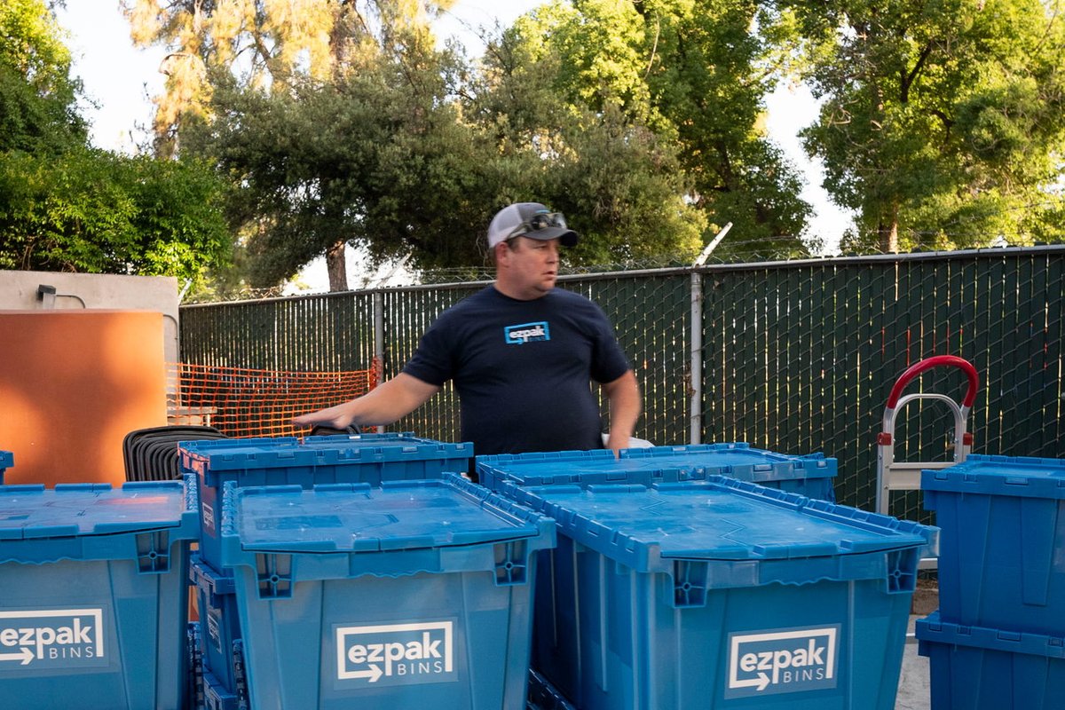 A man in a cap stands behind multiple stacks of blue EZPak Bins moving crates on wheeled dollies in an outdoor lot