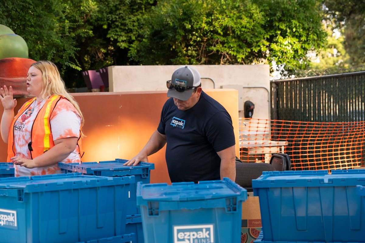 A woman in a safety vest and a man in a branded cap organize multiple stacks of blue EZPak Bins moving crates on wheeled dollies outdoors