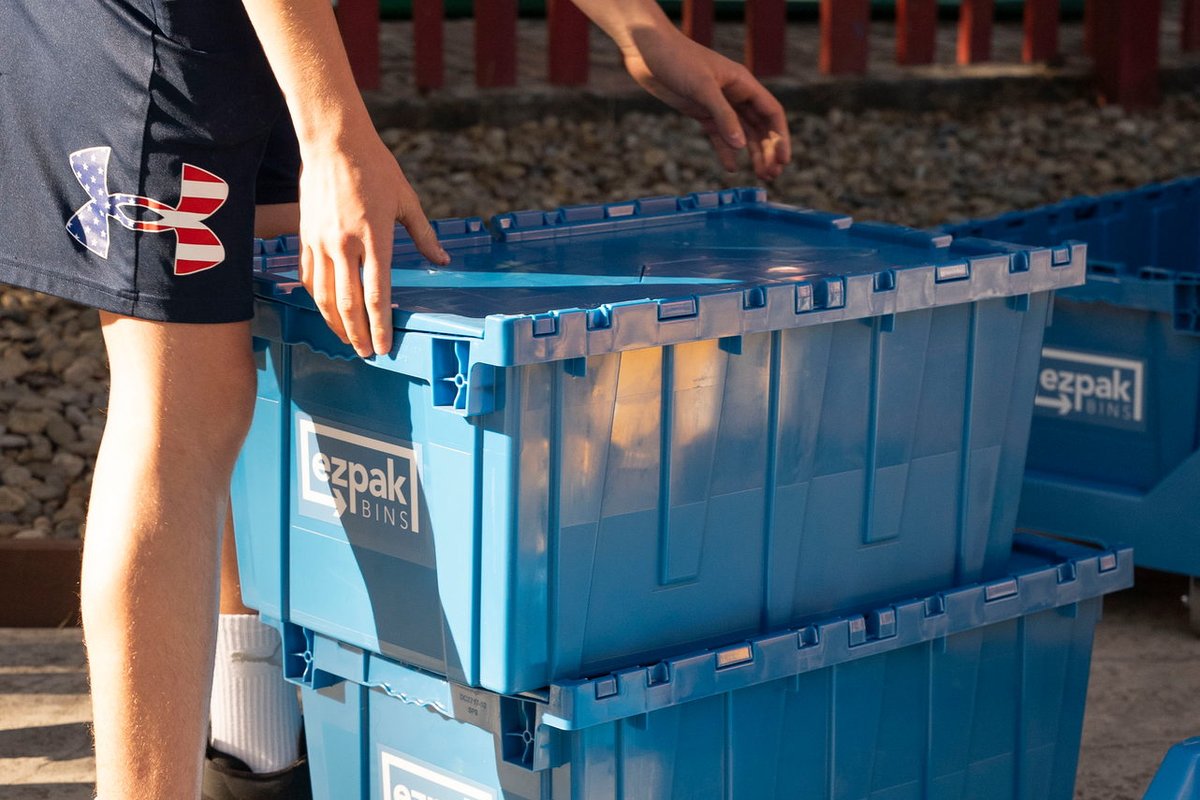 A person closes the lid of stacked blue EZPak Bins moving crates outdoors by a red fence