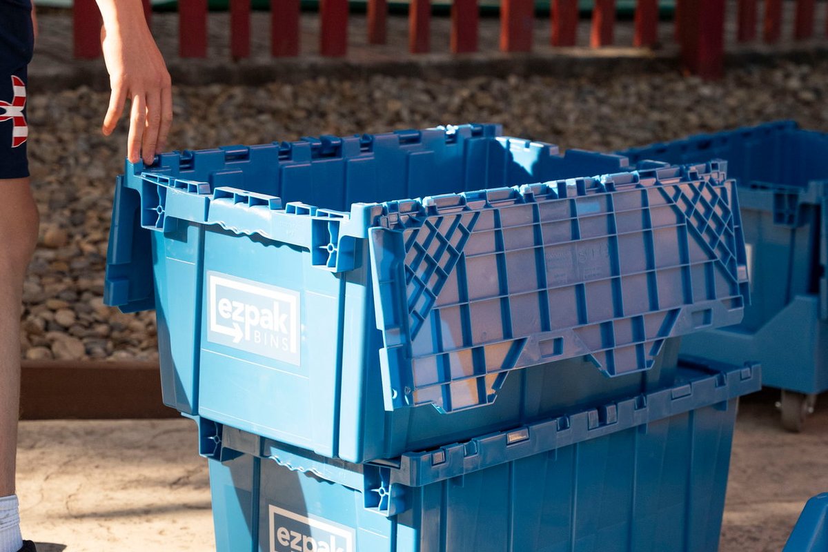 A close-up of stacked open blue EZPak Bins moving crates with a person's hand resting on the top bin, outdoors by a red fence
