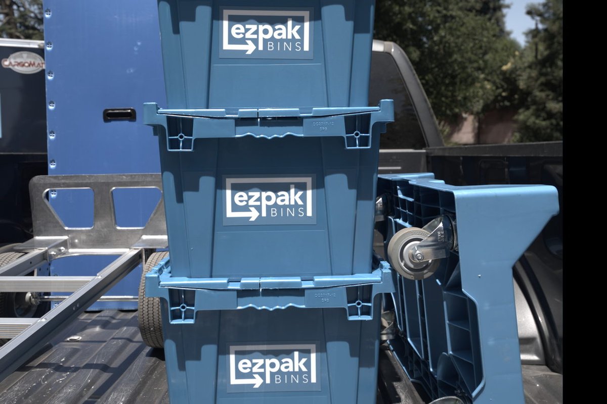 Three blue EZPak Bins moving crates with visible logos stacked together next to a hand truck in the bed of a pickup truck, trees in the background