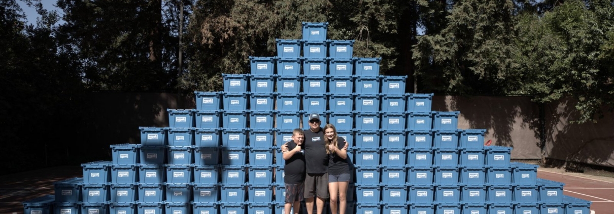 A man, a boy, and a girl pose together in front of a large pyramid-shaped stack of blue EZPak Bins moving crates on an outdoor court