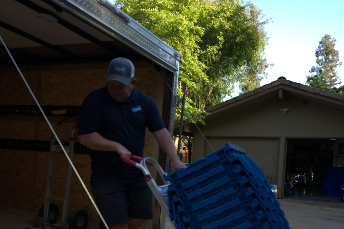 A man wheels a tall stack of blue EZPak Bins moving bins on a hand truck near the open back of a cargo trailer in a driveway