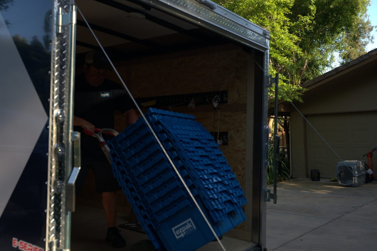 A worker pushes a tall stack of blue EZPak Bins moving bins up into a cargo trailer on a hand truck