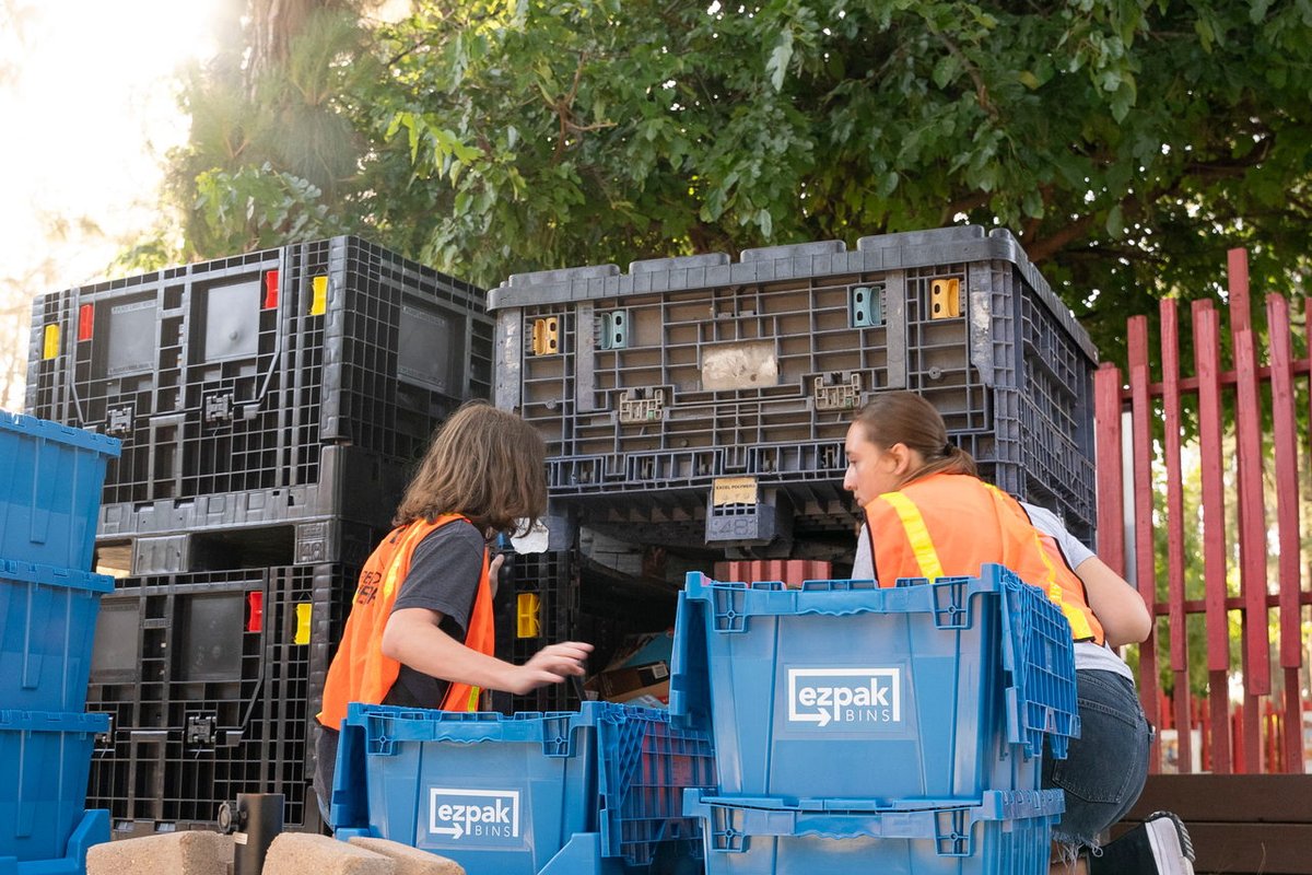 Two people kneel beside blue EZPak Bins moving crates while sorting supplies outdoors
