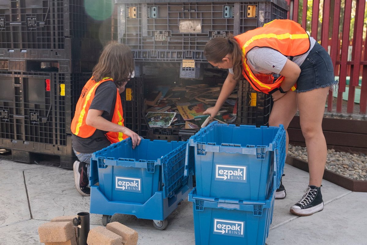 Two workers in orange safety vests bend over to load blue EZPak Bins moving crates onto a dolly near large black storage pallets outdoors under a tree