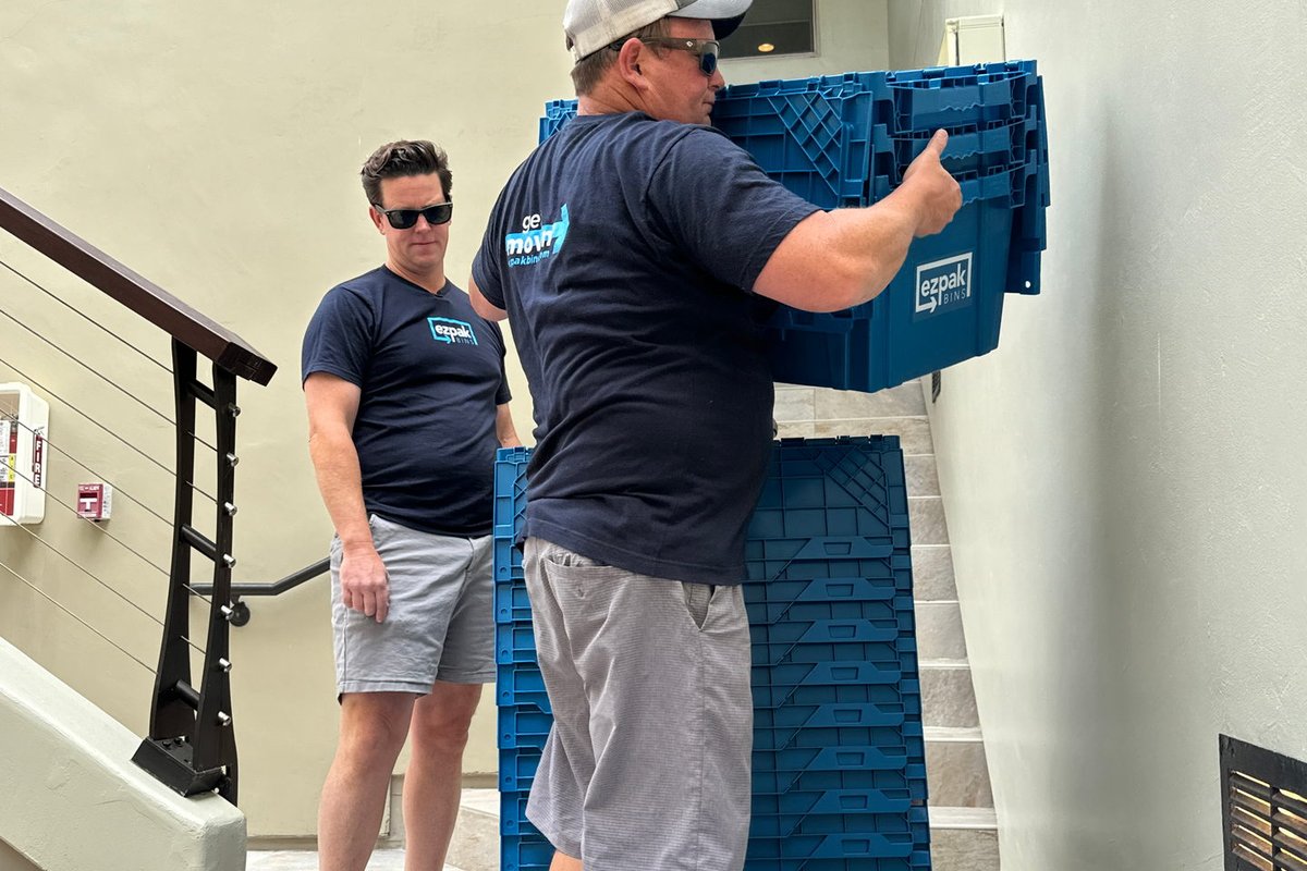 Two men in EZPak Bins shirts stack blue moving bins in a condo stairwell with a wheeled bin dolly in the foreground