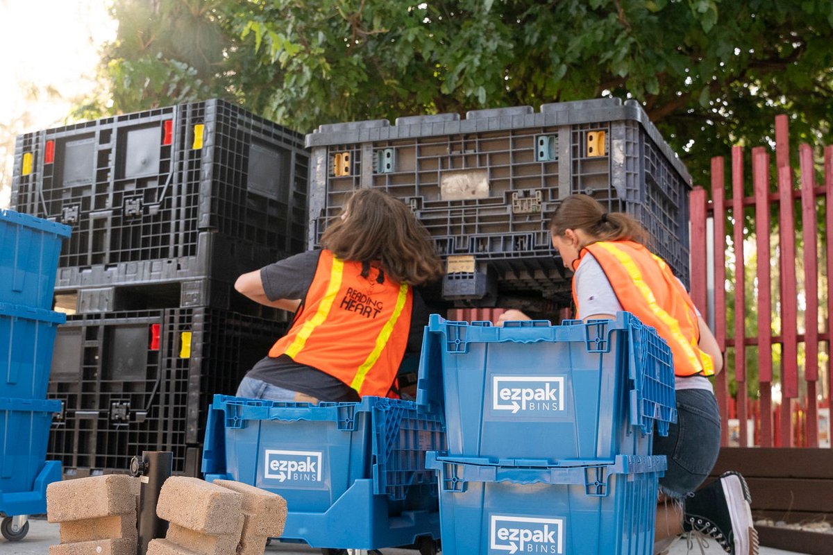 Two workers in safety vests reach into a large storage pallet beside blue EZPak Bins moving crates on an outdoor patio